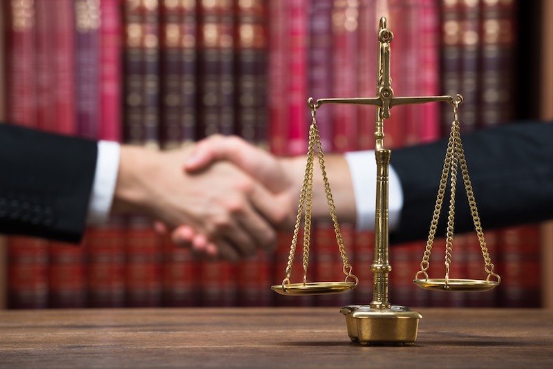 Two People In Suits Shaking Hands Behind A Law Scales Statue