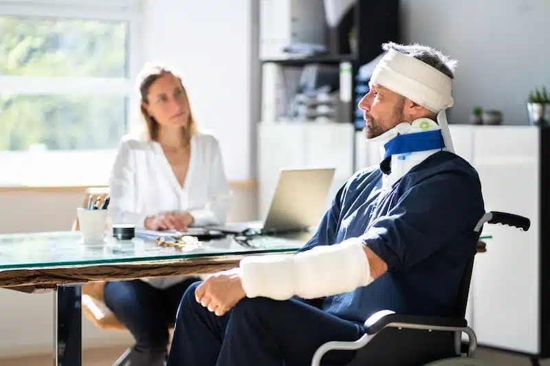 Man In A Wheelchair With Casts And Bandages Talking With A LAwyer
