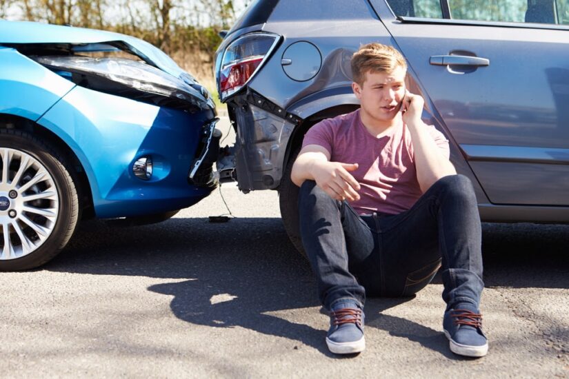 Man Sitting With His Back Against A Damaged Car And Calling Someone